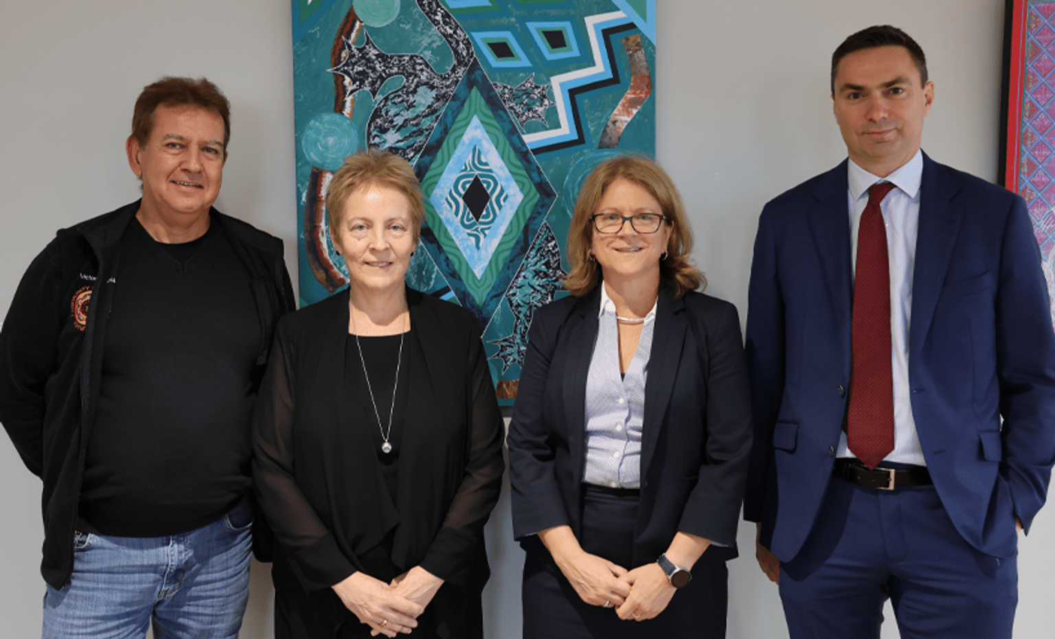 A group shot of VLA representatives standing in front of an blue and teal coloured Aboriginal artwork at the Yoorrook Justice Commission. Lawrence, Louise, Joanna and Dan are in business clothes and smiling towards the camera.