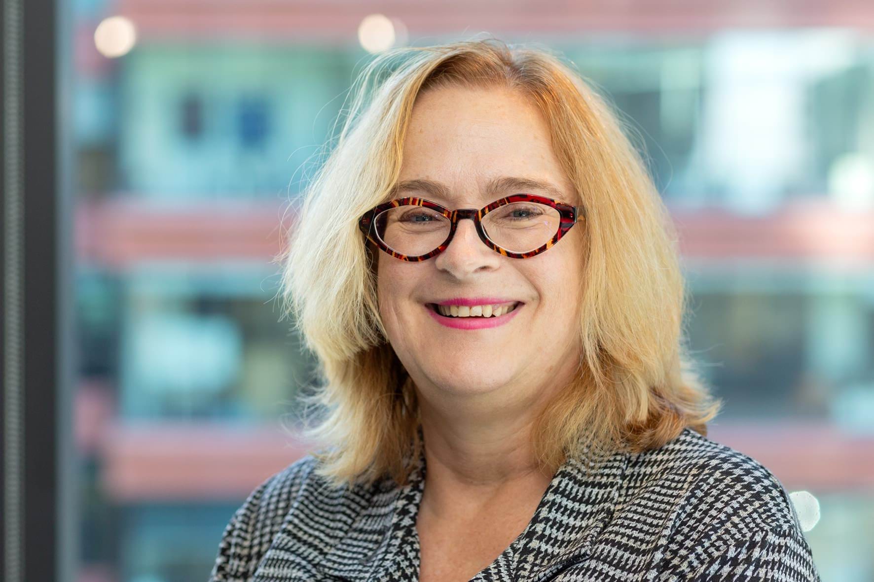 Profile photo of woman with long blonde hair and glasses smiling at camera.