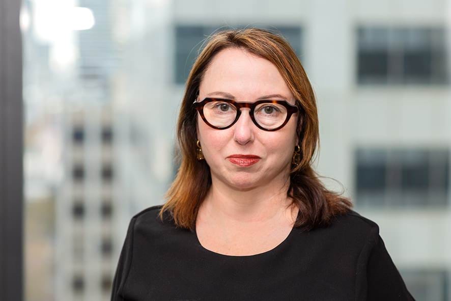 Profile photo of woman with red hair and glasses wearing a black top and looking at camera.